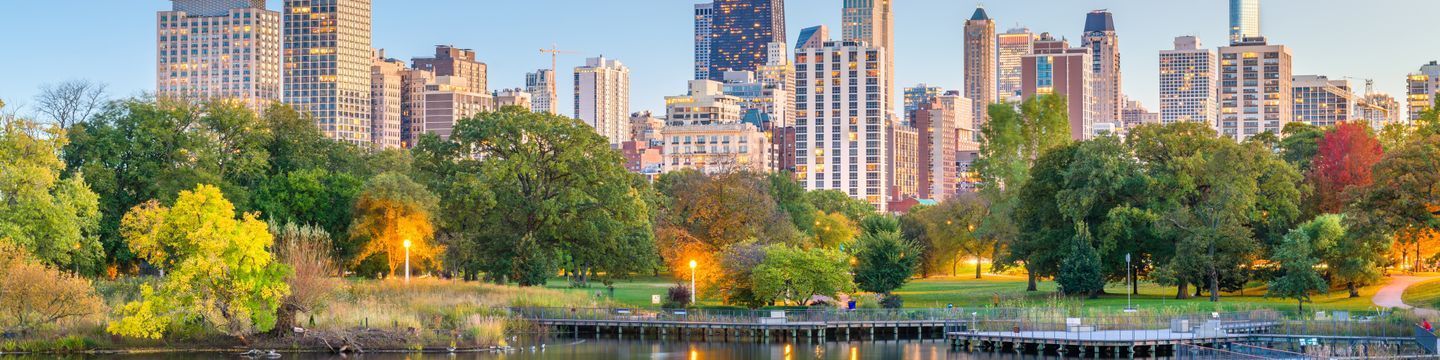 Panoramic view of an Illinois city skyline at dusk with modern high-rise buildings, a foreground of lush green parkland, trees with autumn colors, and a serene pond.