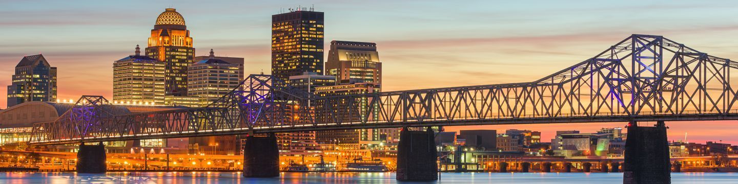 Panoramic view of a Kentucky city skyline at sunset featuring a distinct bridge with a geometric truss design and multiple skyscrapers, illuminated against a colorful sky.