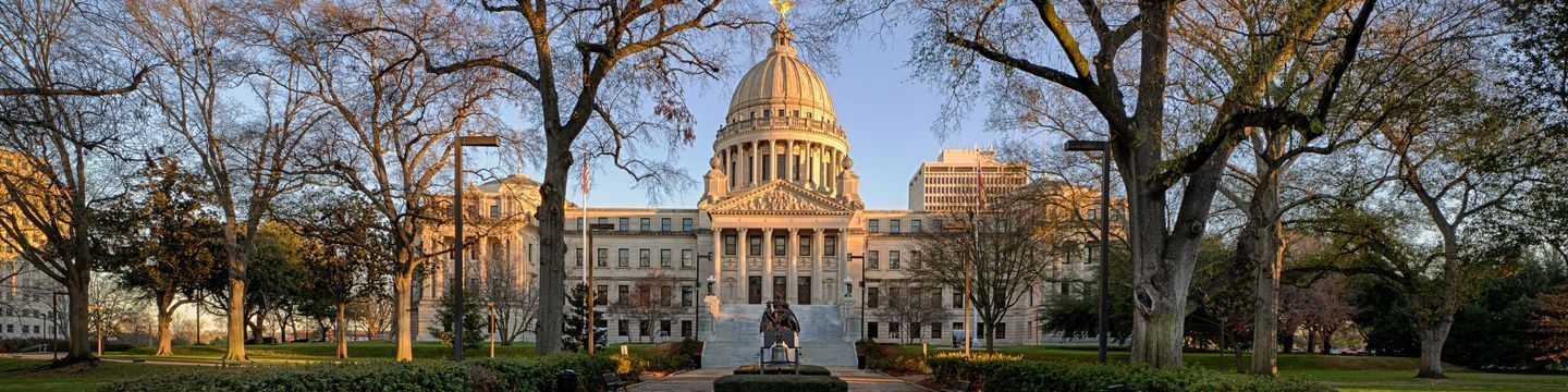Panoramic view of Mississippi's classical domed capitol building framed by towering trees, with a statue in the foreground, under a soft, golden sunset light.
