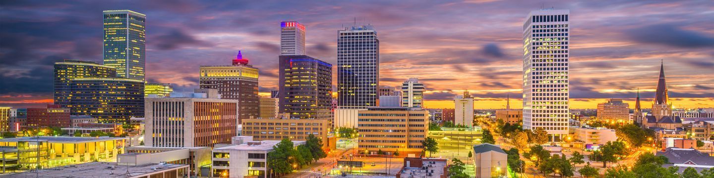 Panoramic view of Oklahoma City's skyline at dusk, showcasing illuminated high-rise buildings under a vibrant sky with clouds.