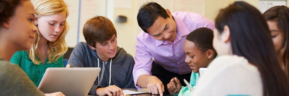 A diverse group of students sitting around a table, looking at a laptop together as a teacher assists them in a classroom setting.
