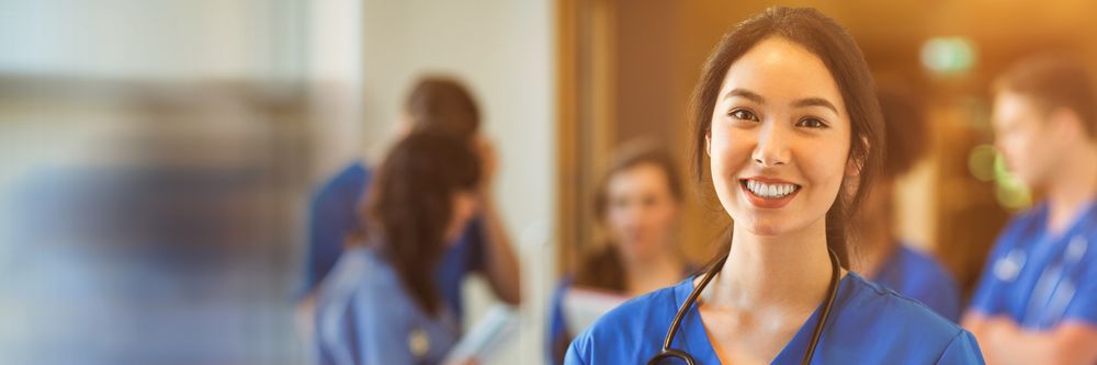 A smiling Asian female healthcare worker in blue scrubs with a stethoscope around her neck, standing in a hospital hallway with blurred colleagues in the background.